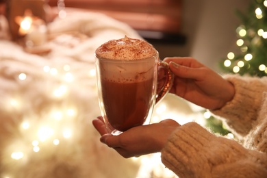 Woman holding cup of delicious drink with whipped cream indoors, closeup. Christmas celebration