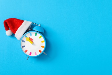 Alarm clock in Santa hat on light blue background, top view with space for text. New Year countdown