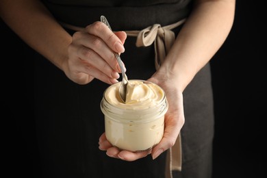 Photo of Woman holding spoon and jar of delicious mayonnaise on black background, closeup