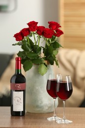 Bottle, glasses of red wine and vase with roses on wooden table in room. Romantic date