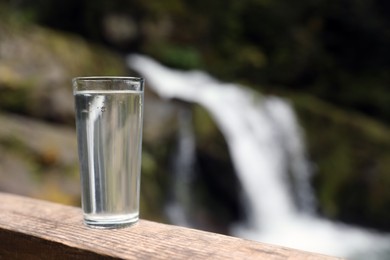 Glass of water on wooden surface near waterfall outdoors. Space for text