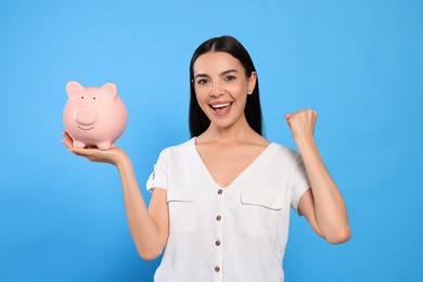 Emotional young woman with piggy bank on light blue background