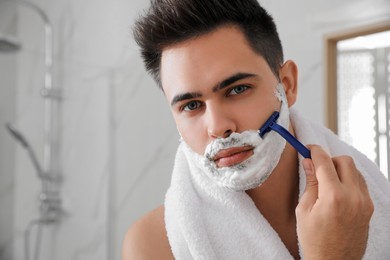Handsome young man shaving with razor in bathroom, space for text