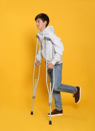 Teenage boy with injured leg using crutches on yellow background