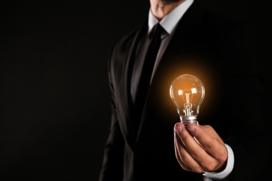 Businessman holding lamp bulb against dark background, closeup. Space for text