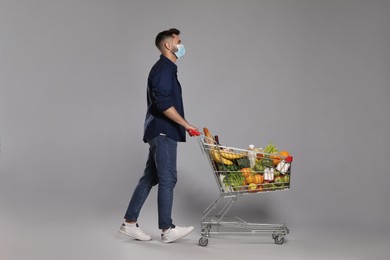 Man with protective mask and shopping cart full of groceries on light grey background