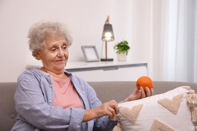 Senior woman finding orange on sofa home. Age-related memory impairment
