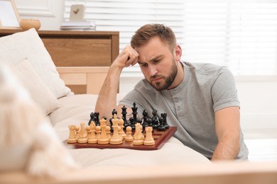 Thoughtful young man playing chess alone on sofa at home