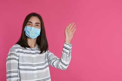 Young woman in protective mask showing hello gesture on pink background, space for text. Keeping social distance during coronavirus pandemic