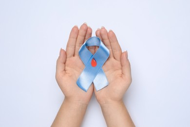 Photo of Woman holding light blue ribbon with drop of blood on white background, top view. Diabetes awareness