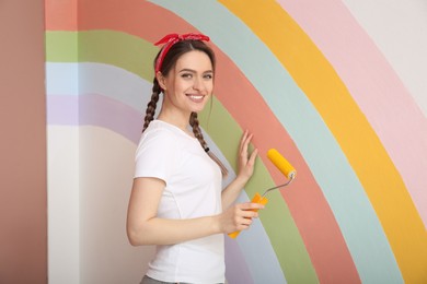 Young woman holding roller near wall with painted rainbow indoors