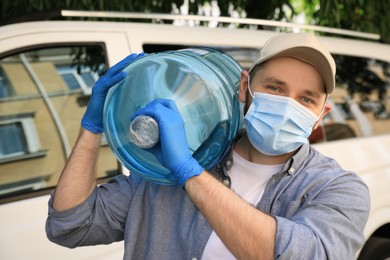 Courier in medical mask holding bottle of cooler water near car outdoors. Delivery during coronavirus quarantine