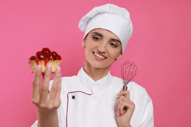 Photo of Happy professional confectioner in uniform with delicious tartlet and whisk on pink background