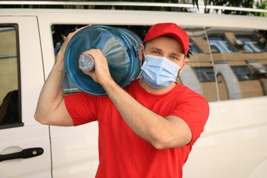 Courier in medical mask holding bottle of cooler water near car outdoors. Delivery during coronavirus quarantine