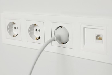 Power sockets with inserted plug on white wall, closeup. Electrical supply
