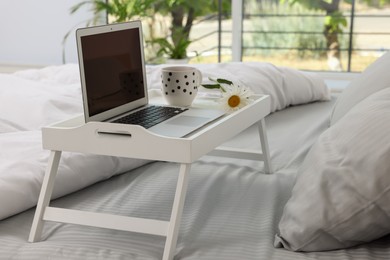 White tray table with laptop, cup of drink and daisy on bed indoors