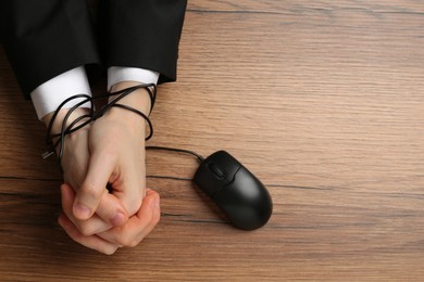 Photo of Man showing hands tied with computer mouse cable at wooden table, top view. Internet addiction