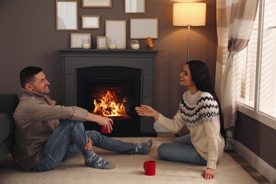 Lovely couple with cups of hot drink spending time together near fireplace at home