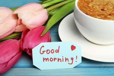 Cup of aromatic coffee, beautiful pink tulips and Good Morning note on light blue wooden table, closeup