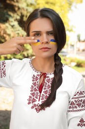 Photo of Sad young woman with drawings of Ukrainian flag on face in park