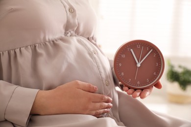 Young pregnant woman holding clock near her belly at home, closeup. Time to give birth