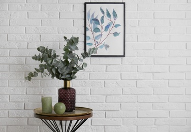 Eucalyptus branches and candles on table near white brick wall with picture. Space for text