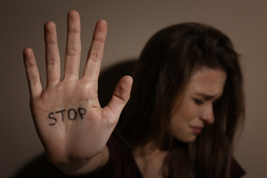 Abused young woman showing palm with word STOP near beige wall, focus on hand. Domestic violence concept
