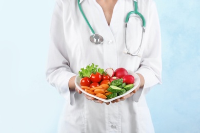 Female doctor holding plate with fresh vegetables on light background. Cardiac diet