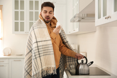 Sick young man putting heating kettle in kitchen. Influenza virus