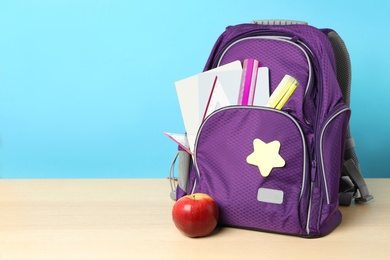 Stylish backpack with different school stationery and apple on wooden table against light blue background, space for text. Back to school