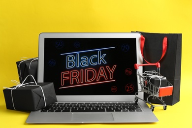 Laptop with Black Friday announcement, small shopping cart and gifts on yellow background
