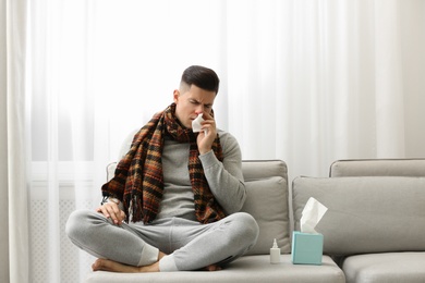 Ill man sitting near nasal spray and box of paper tissues on sofa at home