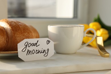 Photo of Cup of coffee, croissant and card with phrase GOOD MORNING near window