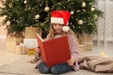 Photo of Cute little girl in Santa hat reading book near tree and gifts at home. Christmas atmosphere
