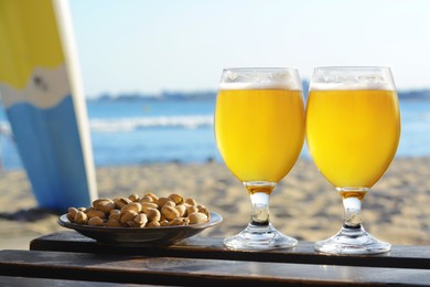 Glasses of cold beer and pistachios on wooden table near sea, space for text