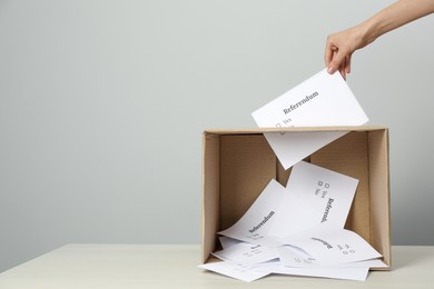 Woman putting referendum ballot in box on table against light grey background, closeup. Space for text