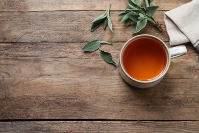 Cup of aromatic sage tea and fresh leaves on wooden table, above view. Space for text