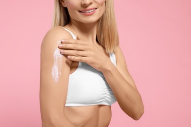 Photo of Woman applying body cream onto her arm against pink background, closeup. Space for text