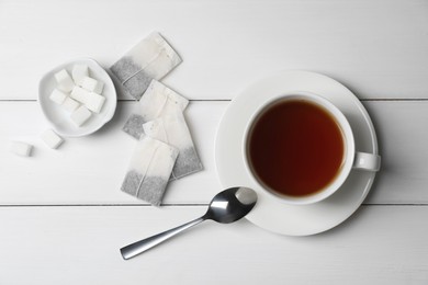 Tea bags and sugar near cup of hot drink on white wooden table, flat lay
