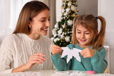 Happy mother and daughter making paper snowflake at table near Christmas tree indoors