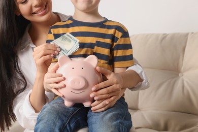 Mother and her son putting money into piggy bank at home, closeup
