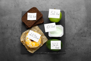 Food products with calorific value tags on dark grey table, flat lay. Weight loss concept