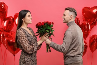 Photo of Boyfriend presenting bouquet of roses to his girlfriend near heart shaped air balloons on red background. Valentine's day celebration
