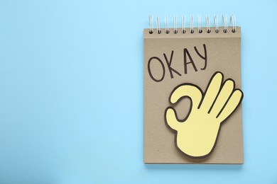 Photo of Notepad with word Okay and paper cutout of OK hand gesture on light blue background, top view. Space for text