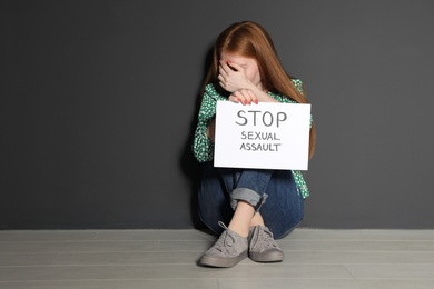 Young woman holding card with words STOP SEXUAL ASSAULT while sitting near grey wall. Space for text