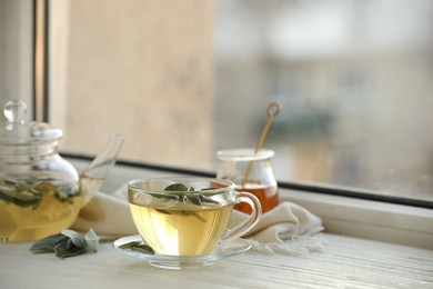 Photo of Cup of sage tea and green leaves on white wooden windowsill. Space for text
