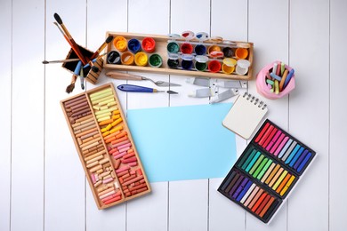 Blank sheet of paper, colorful chalk pastels and other drawing tools on white wooden table, flat lay. Modern artist's workplace