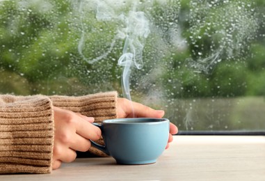 Woman with cup of hot drink at wooden table near window on rainy day, closeup