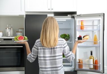 Photo of Choice concept. Woman holding plates with fruits and sweets near refrigerator in kitchen, back view