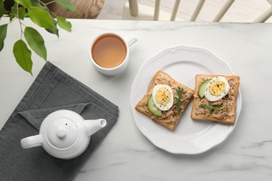 Photo of Delicious sandwiches with tuna, vegetables, boiled egg and cup of coffee on white marble table indoors, flat lay. Tasty breakfast